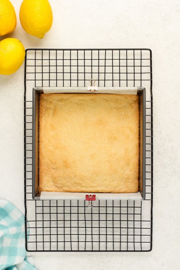 Baked shortbread crust rests in a lined baking pan on a black wire cooling rack. Three lemons are placed in the top left corner. A folded blue and white checkered cloth is at the bottom left.