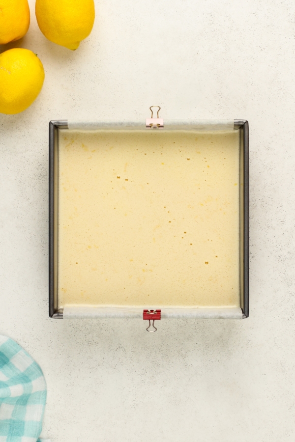 A square baking pan filled with uncooked lemon batter lined with parchment paper and secured by binder clips. Three whole lemons are in the top left corner, and a blue and white checkered cloth is partially visible at the bottom left.