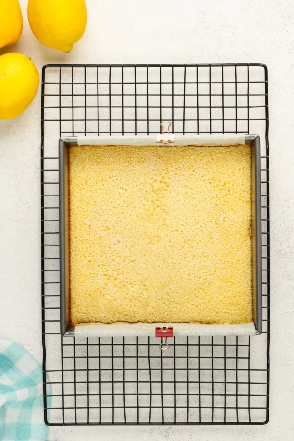 A square pan filled with freshly baked lemon bars cooling on a black wire rack. The cake sits in a square pan with parchment paper clipped on the sides. Two whole lemons are placed in the top left corner.