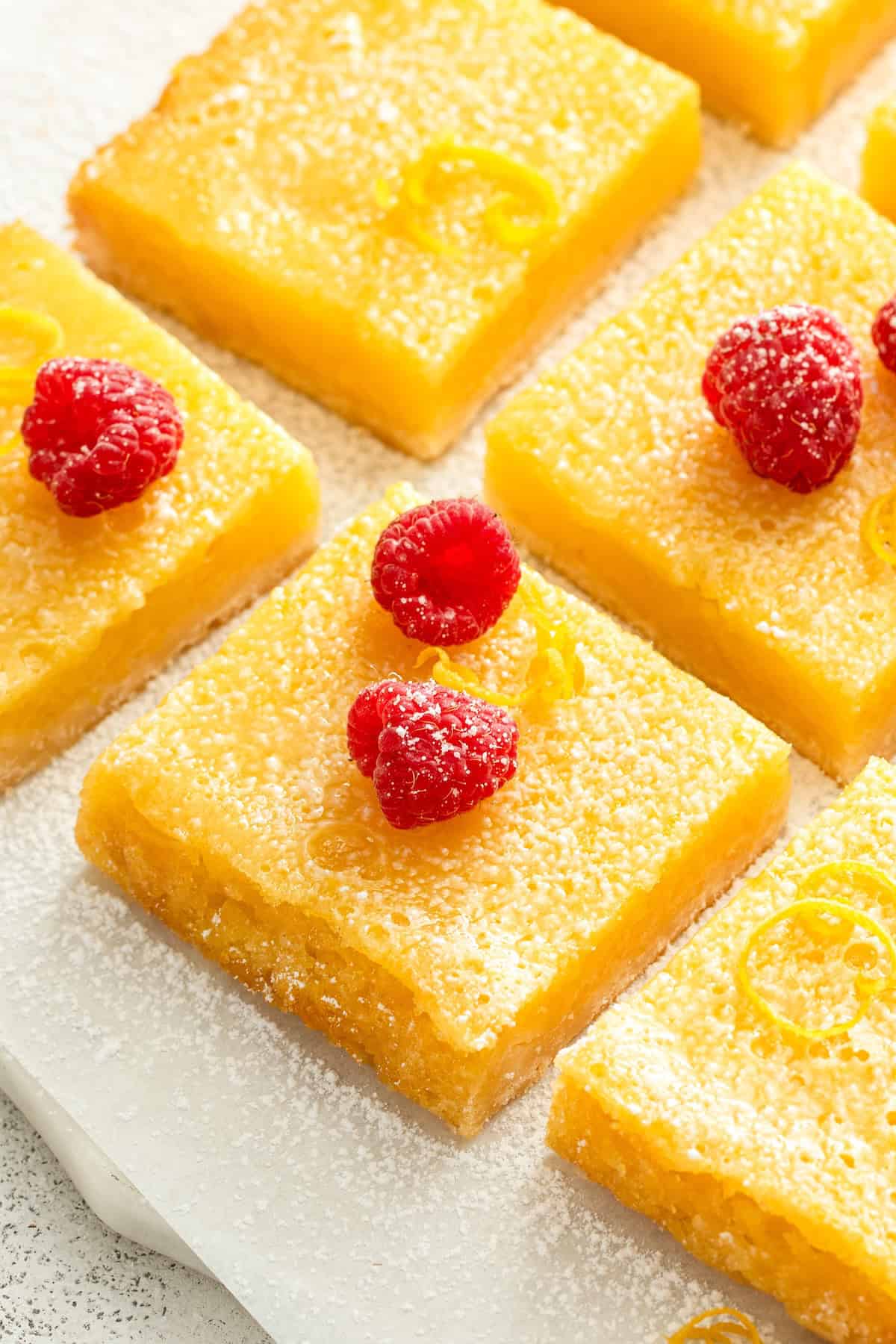 Slices of lemon bars dusted with powdered sugar are arranged on a white surface. They are garnished with fresh raspberries and lemon zest.
