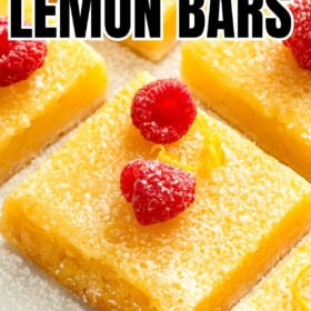 Close-up of lemon bars topped with powdered sugar and raspberries. The bars are cut into squares and set on a light-colored surface.