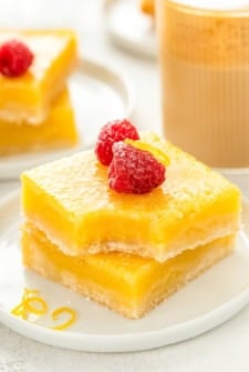 A stack of two lemon bars on a white plate, topped with powdered sugar, fresh raspberries, and lemon zest. The top lemon bar has a bite taken out of it.