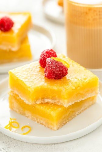 A stack of two lemon bars on a white plate, topped with powdered sugar, fresh raspberries, and lemon zest. The top lemon bar has a bite taken out of it.