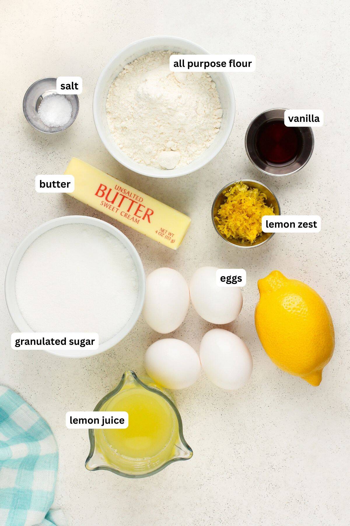 Ingredients for baking this easy lemon bars recipe are arranged on a beige surface. They include all-purpose flour, a stick of unsalted butter, granulated sugar, eggs, lemon zest, a lemon, lemon juice, vanilla, and salt, with labels for each item. A teal cloth is in the corner.