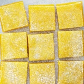 Nine lemon bars dusted with powdered sugar are arranged in a grid on a sheet of parchment paper atop a wire cooling rack, set on a light gray textured surface.