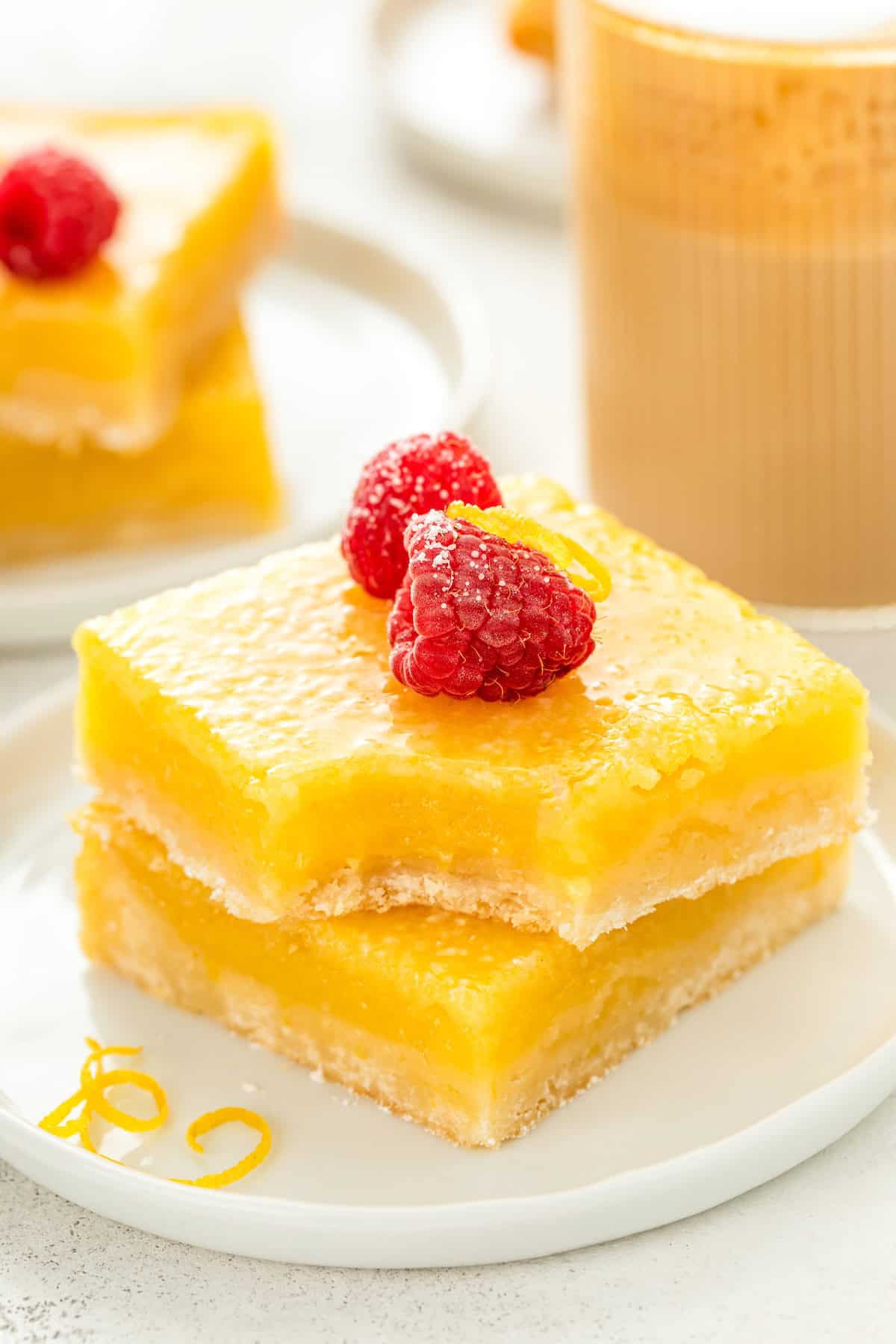 Made with just 7 simple ingredients, this easy Lemon Bars recipe is tangy and sweet with a buttery shortbread crust that melts in your mouth! A stack of two lemon bars on a white plate, topped with powdered sugar, fresh raspberries, and lemon zest. The top lemon bar has a bite taken out of it.