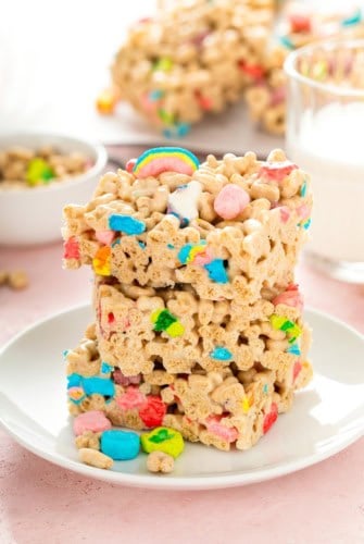 Three stacked Lucky Charms rice krispie treats with colorful marshmallows sit on a white plate. A bowl of cereal and a glass of milk are in the background on a light pink table.