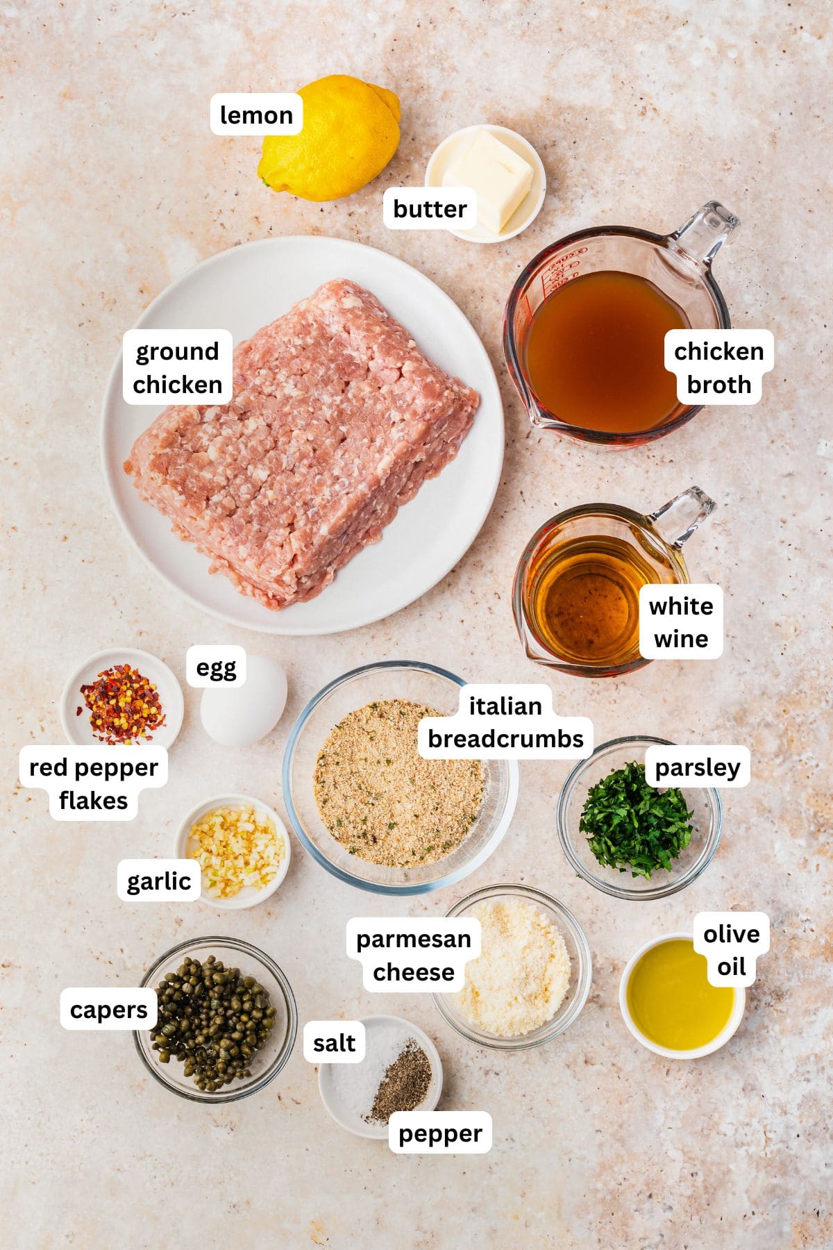 A flat lay of chicken piccata meatball recipe ingredients including ground chicken, a lemon, butter, chicken broth, white wine, red pepper flakes, an egg, minced garlic, Italian breadcrumbs, chopped parsley, parmesan cheese, olive oil, capers, salt, and pepper.