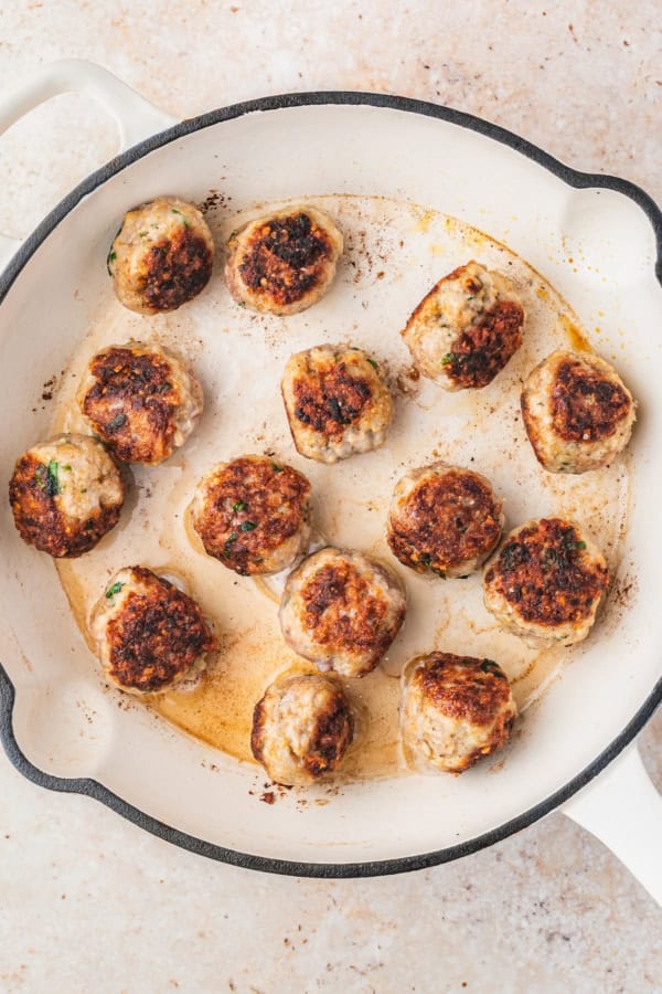 A white skillet with browned chicken meatballs, some with herbs visible, cooking in their juices on a light tan countertop.