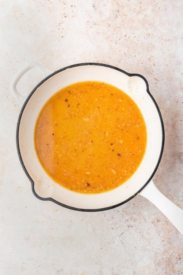 A white pot with a handle contains a yellow-orange lemon sauce with visible small bits of leftover cooked meatball pieces, set on a light-colored textured surface.