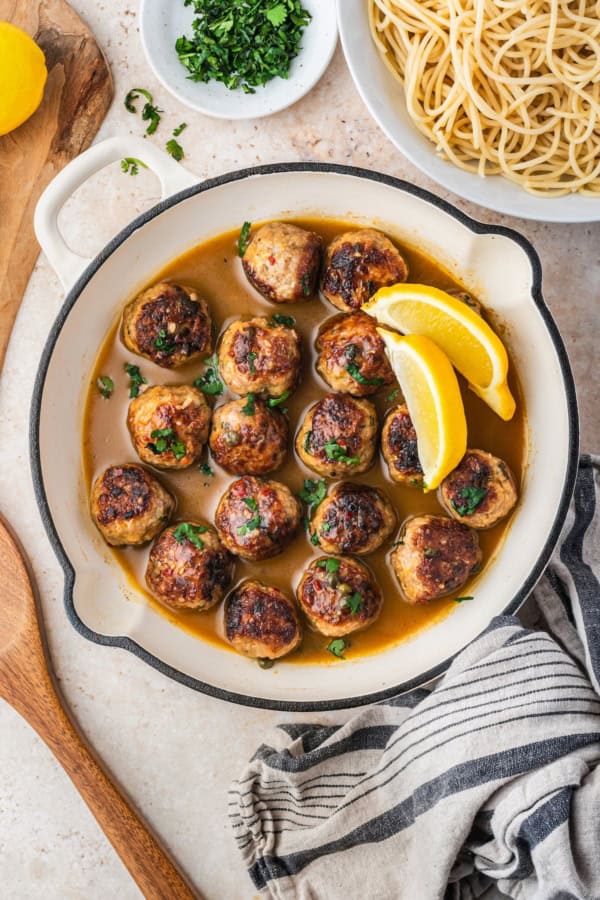 A pan filled with browned chicken meatballs in a lemon-caper sauce, garnished with chopped herbs and lemon wedges, sits next to a bowl of cooked spaghetti, a small plate of herbs, and a striped kitchen towel.