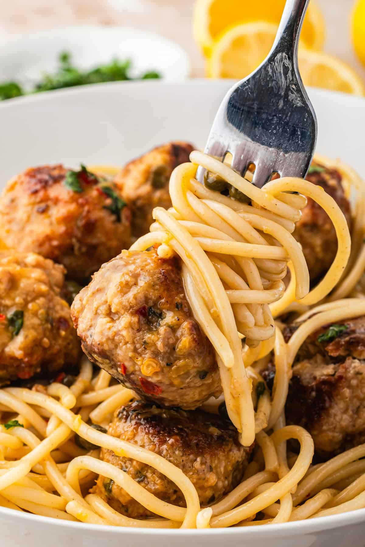 A fork twirls spaghetti noodles above a bowl filled with spaghetti and several chicken piccata meatballs, garnished with fresh herbs. In the background, there are slices of lemon and a plate with chopped parsley.