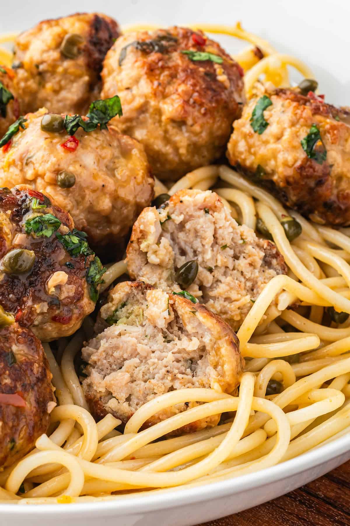 A close-up of spaghetti topped with several browned chicken meatballs, one of which is cut open to show the inside, garnished with herbs and capers.