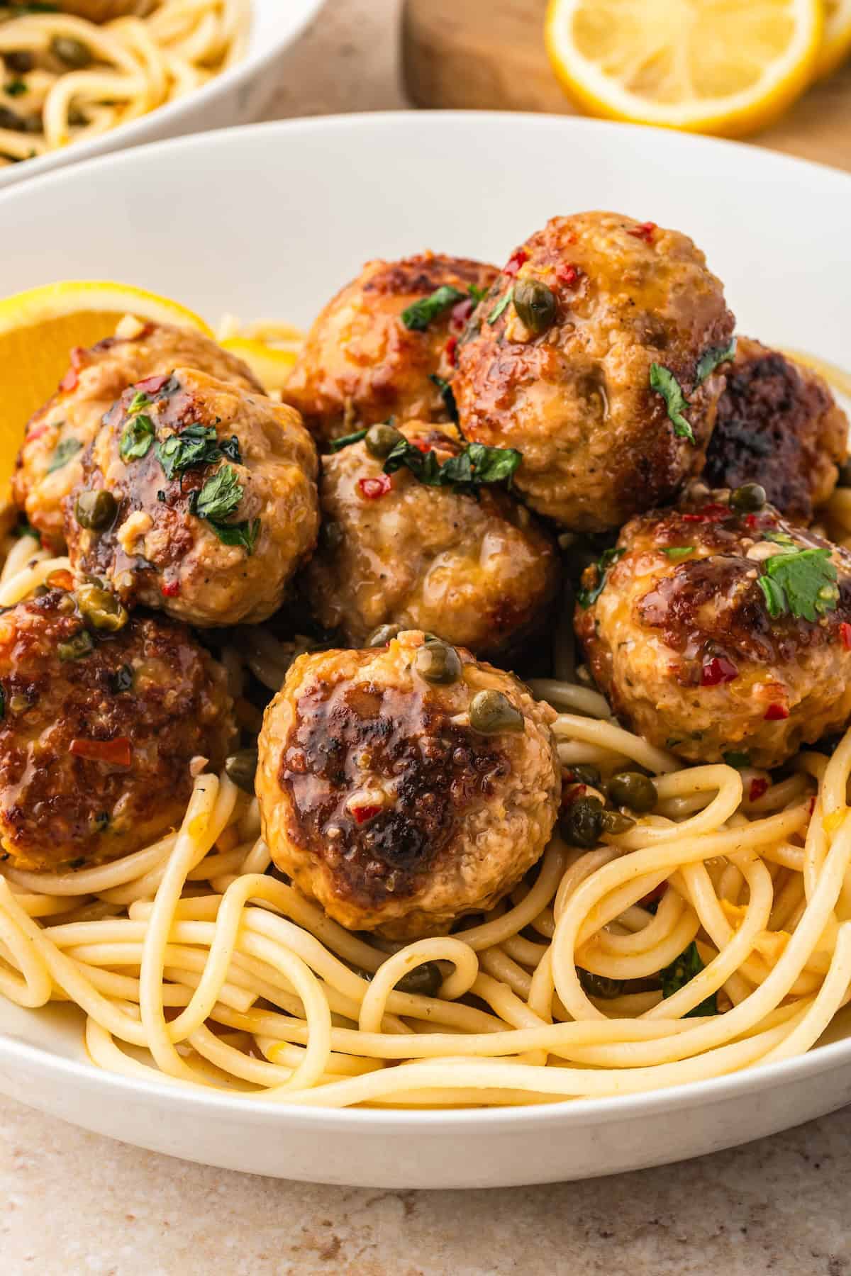 A white bowl filled with spaghetti topped with several browned chicken piccata meatballs in a lemon sauce with capers, garnished with chopped herbs and with a lemon wedge on the side.