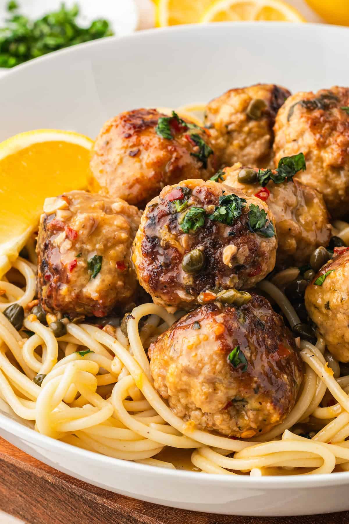A close-up of spaghetti topped with browned chicken piccata meatballs in a lemon caper sauce, garnished with chopped herbs, and served with lemon wedges in a white bowl.