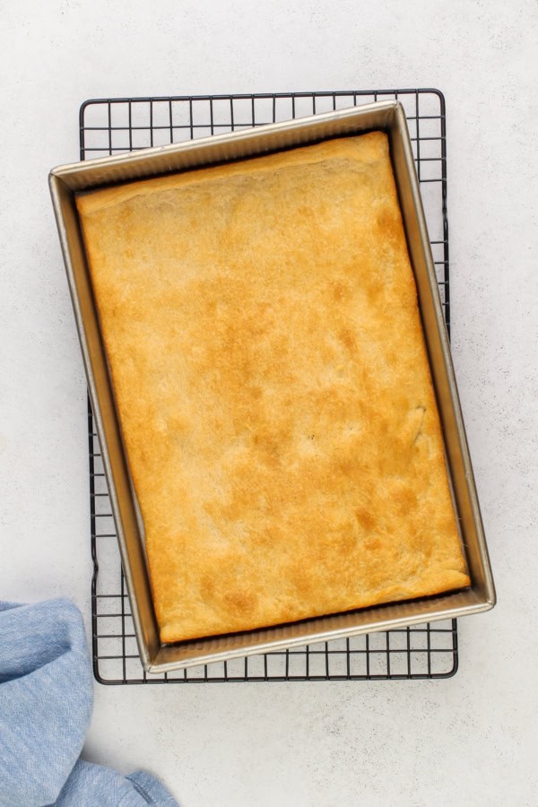 A golden-brown rectangular baked crescent dough sits in a metal baking pan on a cooling rack, with a light blue cloth partially visible in the corner.