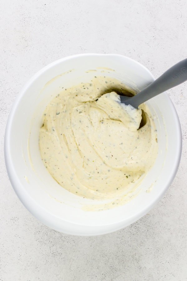 A white bowl filled with cream cheese, sour cream, and ranch seasoning mixture with green herb flecks. A gray spoon is partially submerged in the mixture, resting on the side of the bowl.