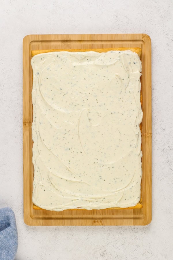 A rectangular baked crescent roll dough topped with a layer of creamy ranch and cream cheese mixture, placed on a wooden cutting board against a light background. A blue cloth is partially visible on the lower left corner.