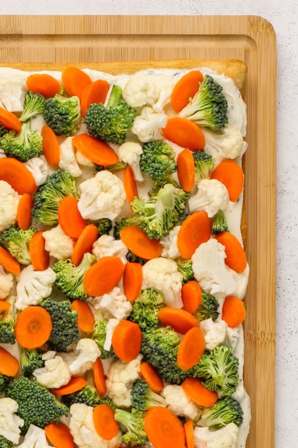 A close-up of a rectangular vegetable pizza with crescent roll dough crust topped with a creamy ranch sauce, sliced carrots, broccoli florets, and cauliflower, displayed on a wooden cutting board.