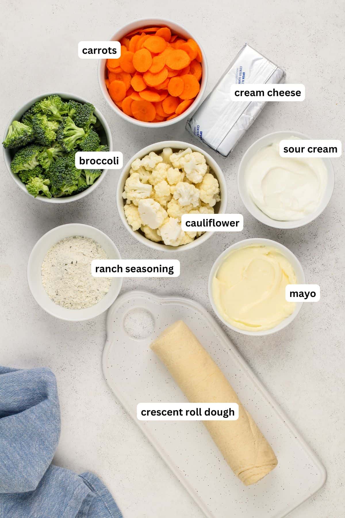 A flat lay of labeled ingredients for this veggie pizza recipe with crescent rolls arranged in bowls: chopped carrots, broccoli, cauliflower, cream cheese, sour cream, mayo, ranch seasoning, and a roll of crescent dough on a white board.