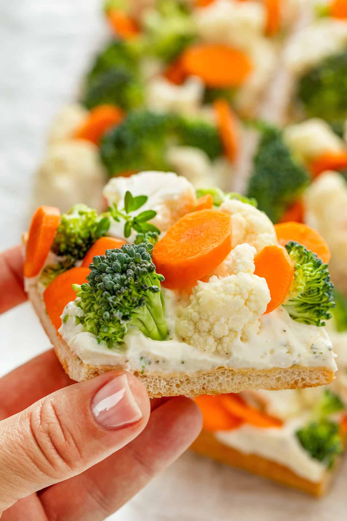 A hand holding a square piece of crescent roll veggie pizza topped with broccoli, cauliflower, and carrot slices over a creamy ranch layer with a soft crust. More veggie pizza pieces are blurred in the background.