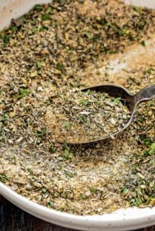 A close-up of a rustic metal spoon scooping up a blend of Greek seasoning made of dried herbs and Greek spices from a bowl.