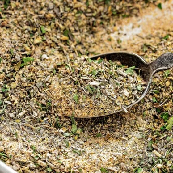 A close-up of a rustic metal spoon scooping up a blend of Greek seasoning made of dried herbs and Greek spices from a bowl.