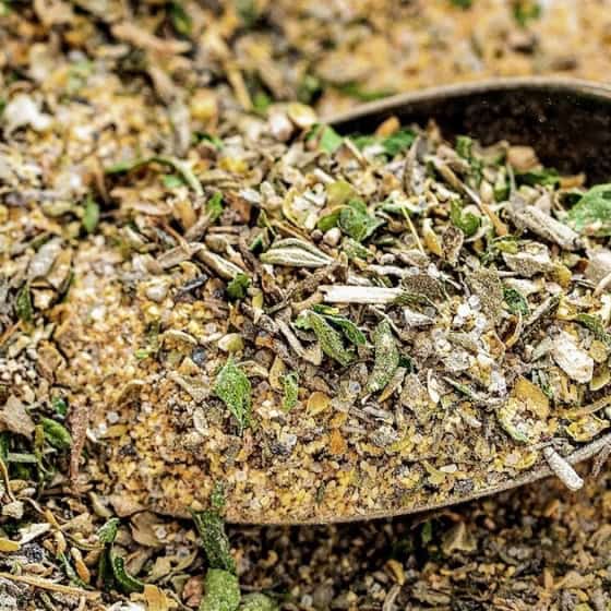 A close-up of a metal spoon filled with a blend of Greek seasoning, with more of the mixture scattered around it.