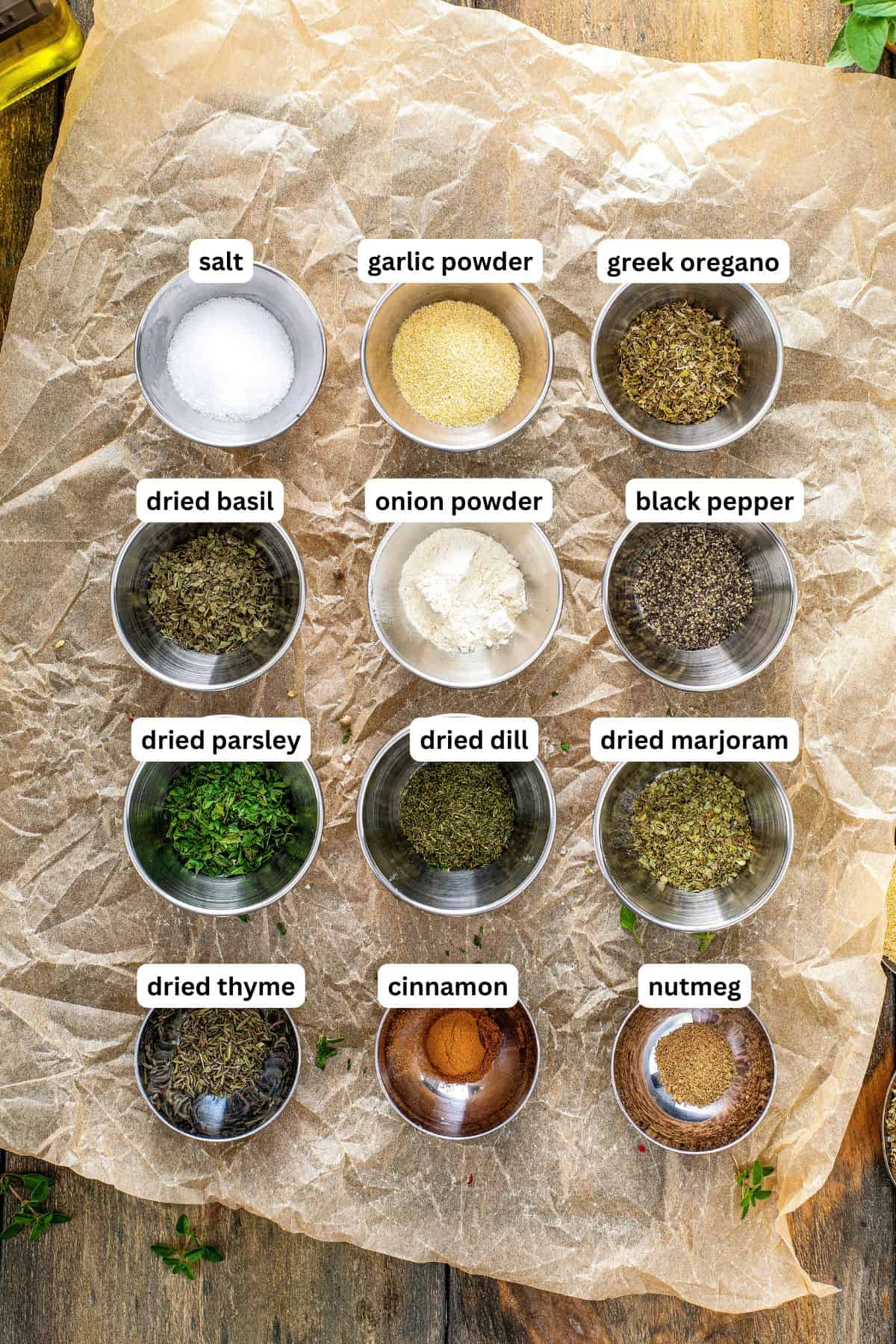 Ingredients for Greek seasoning recipe arranged in a grid on parchment paper. Each bowl is labeled, including salt, garlic powder, greek oregano, dried basil, onion powder, black pepper, dried parsley, dried dill, dried marjoram, dried thyme, cinnamon and nutmeg.