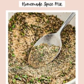 A bowl of homemade Greek seasoning spice mix with a silver spoon, topped with dried herbs.