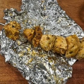Grilled chicken pieces seasoned with greek seasoning on a metal skewer rest on crumpled aluminum foil, placed on a wooden surface. The chicken is seasoned and cooked, with visible grill marks and a slightly charred appearance.