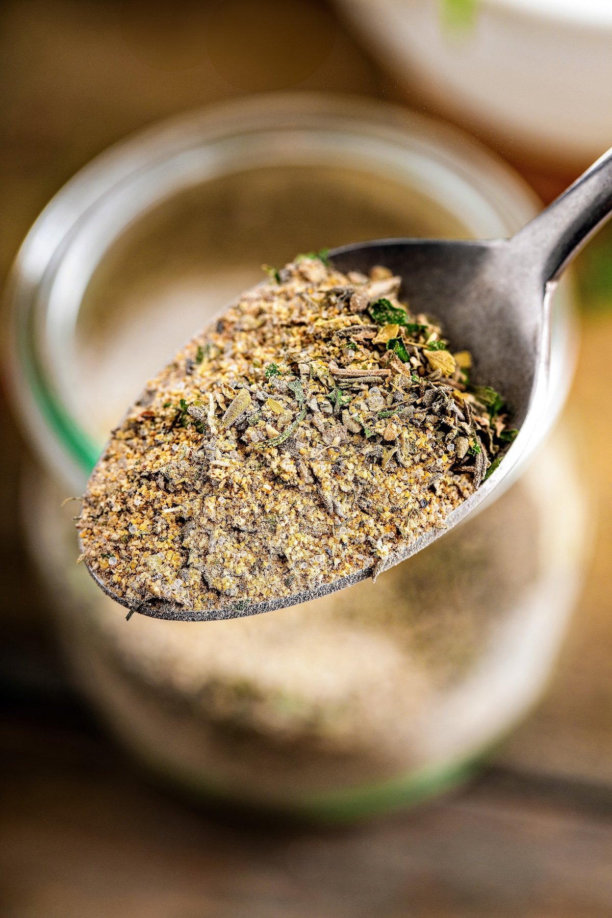 A close-up of a spoonful of dried Greek chicken seasoning with dried herbs and spices held above a glass jar filled with the same mixture.