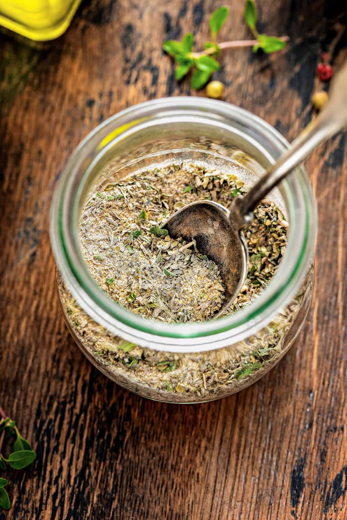 A glass jar filled with mixed dried greek spices sits on a rustic wooden surface, with a metal spoon partially submerged in the herbs. Small green sprigs and a bottle are nearby.
