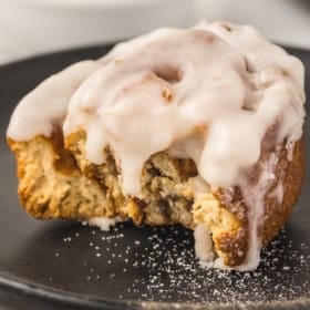 A frosted honey bun topped with white icing sits on a dark plate, with a large bite taken out, revealing its soft, swirled interior.