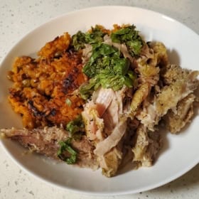A white plate with shredded roast pork pernil topped with chopped herbs, served next to a portion of orange-colored arroz con gandules on a light countertop.