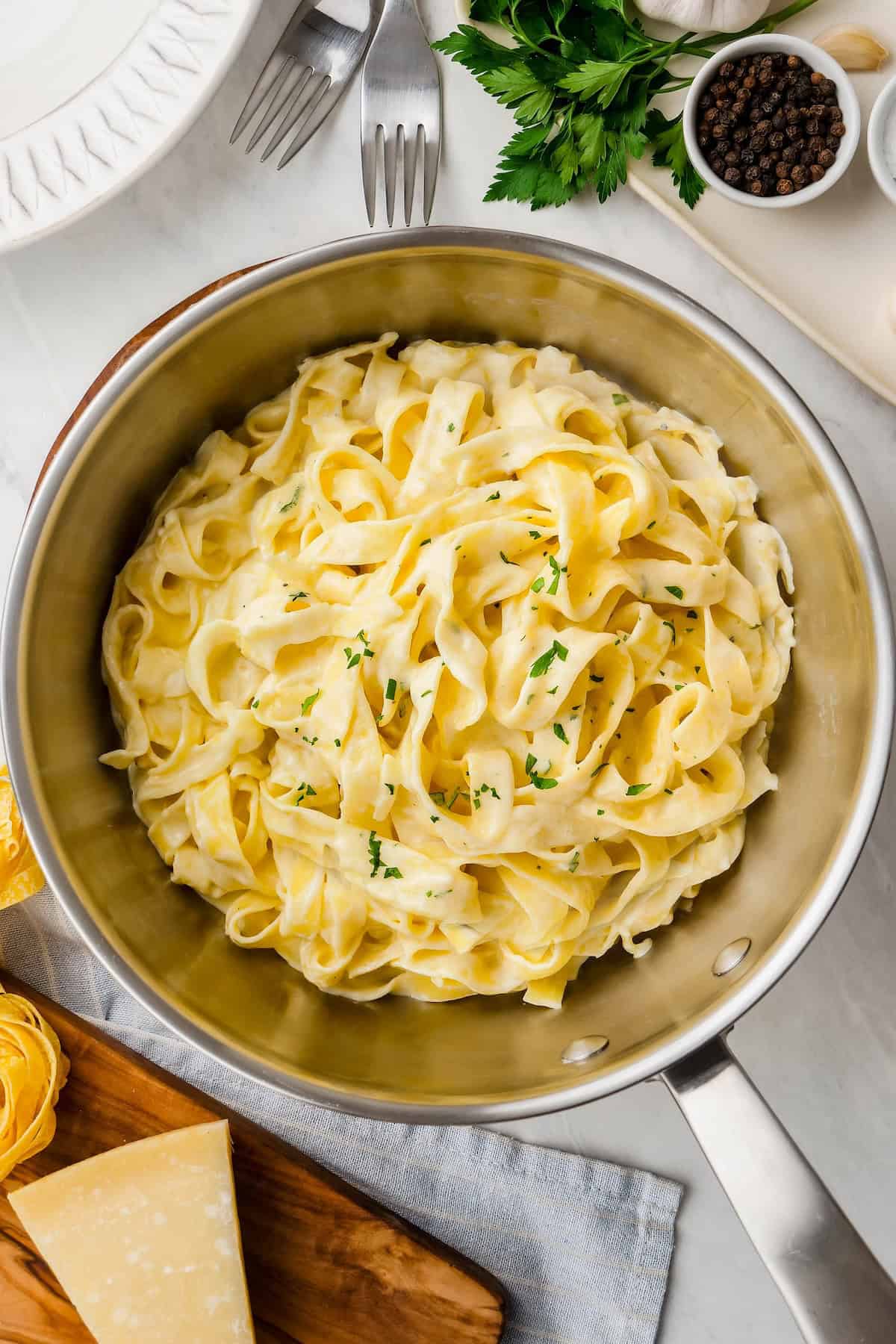 No flour, no cream cheese โ just butter, cream, garlic, and Parmesan in this thick and creamy homemade Alfredo Sauce recipe. Better than any restaurant! A pot filled with creamy homemade Alfredo sauce tossed with fettuccine pasta, garnished with chopped parsley, siting on a white countertop. A wedge of parmesan cheese, uncooked pasta, peppercorns, and fresh herbs surround the pot.