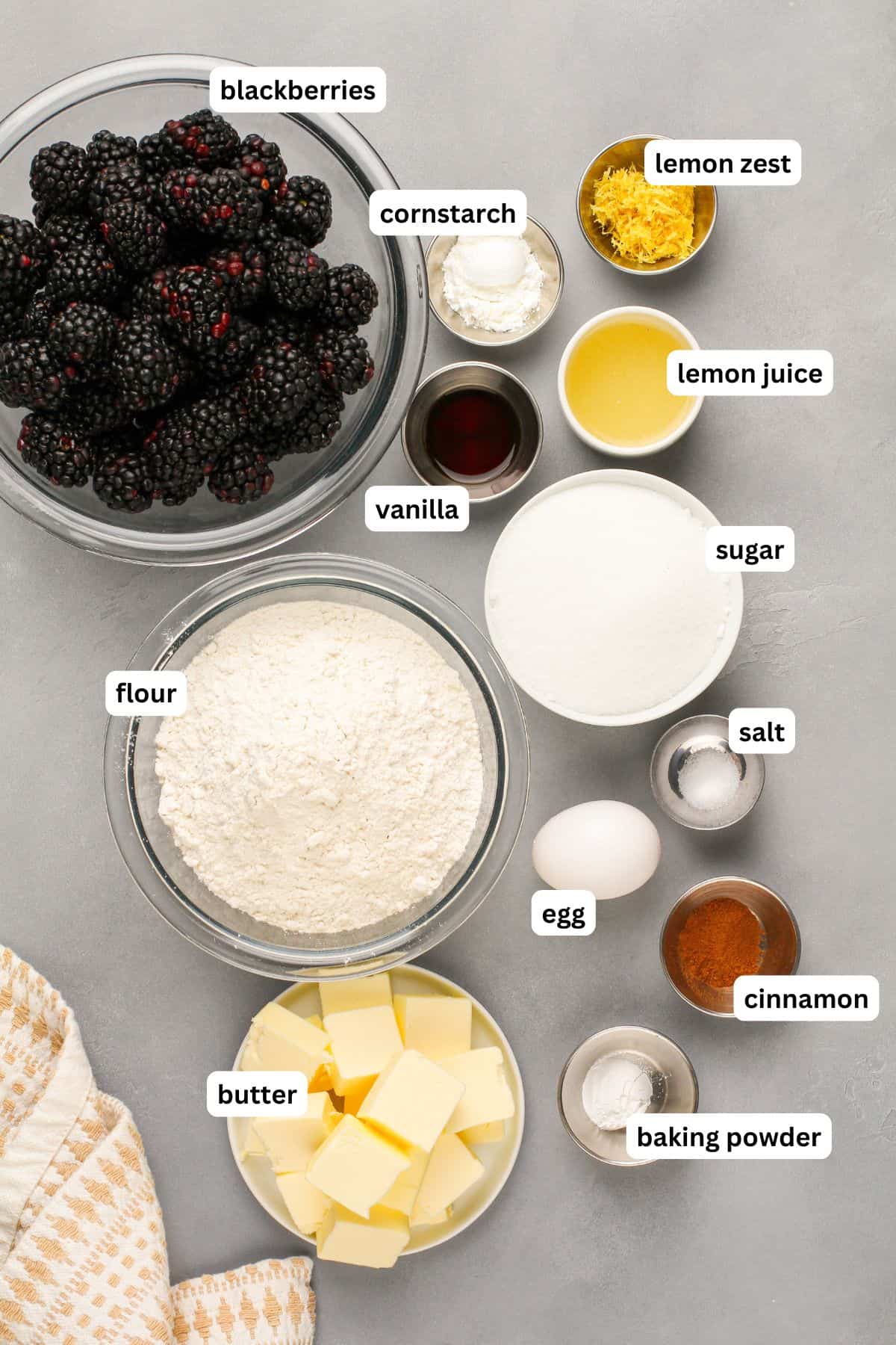 Love pie but hate the hassle? These Blackberry Pie Bars deliver all the flavor of blackberry pie without having to roll out the pie dough! A flat lay of ingredients to make blackberry pie bars recipe arranged in bowls, including blackberries, flour, butter, sugar, an egg, lemon zest, lemon juice, cornstarch, vanilla, salt, cinnamon, and baking powder, all labeled on a gray surface.