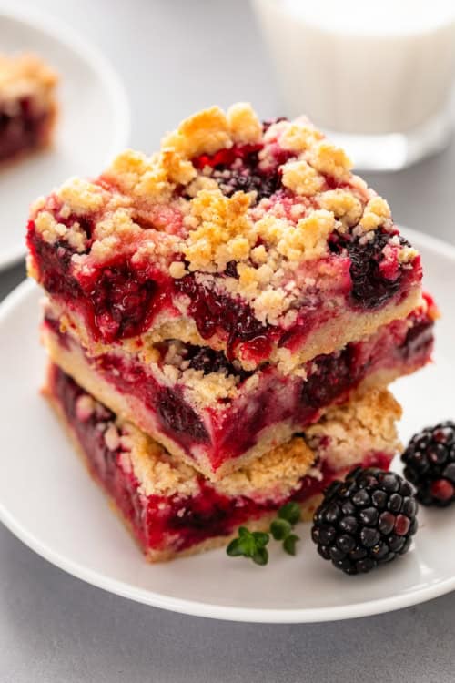 Two crumbly blackberry pie bars stacked on a white plate, filled with blackberries and a golden oat topping, with fresh blackberries and a sprig of thyme next to them. A glass of milk is blurred in the background.