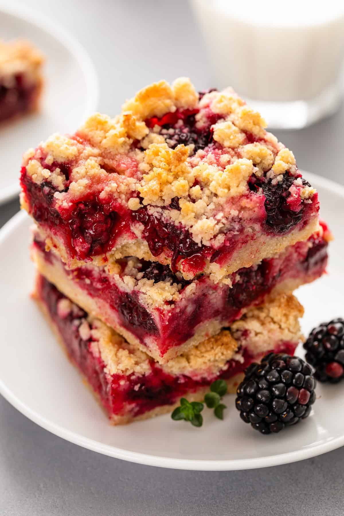 Love pie but hate the hassle? These Blackberry Pie Bars deliver all the flavor of blackberry pie without having to roll out the pie dough! Two crumbly blackberry pie bars stacked on a white plate, filled with blackberries and a golden oat topping, with fresh blackberries and a sprig of thyme next to them. A glass of milk is blurred in the background.