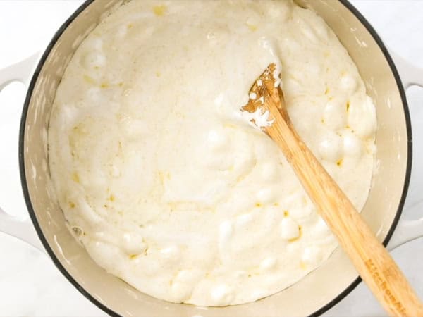 A wooden spoon stirs a creamy, melted marshmallow and brown butter mixture with visible marshmallow lumps in a white pot.