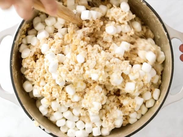 A close-up of a large pot filled with rice cereal and mini marshmallows being mixed together with a wooden spoon.