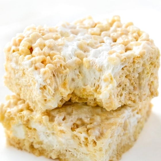 Two gooey Brown Butter Rice Krispie Treats stacked on a white plate, showing crispy cereal bound together with melted marshmallow, with a bite taken out of the top treat.