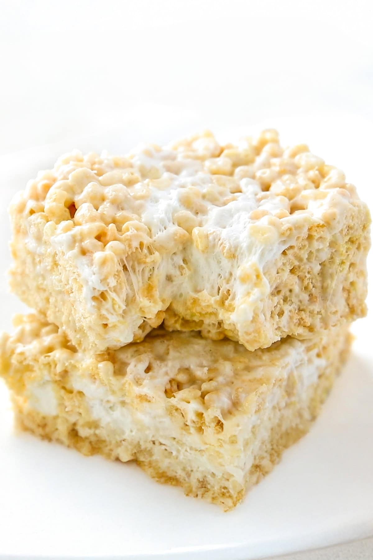 Two gooey Brown Butter Rice Krispie Treats stacked on a white plate, showing crispy cereal bound together with melted marshmallow, with a bite taken out of the top treat.