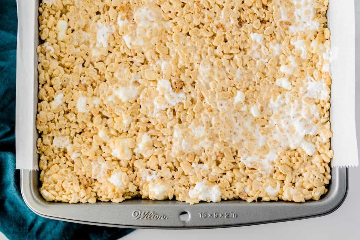 A baking pan filled with brown butter Rice Krispie treats, showing a golden, crispy surface with visible melted marshmallows and a piece of parchment paper lining the side.