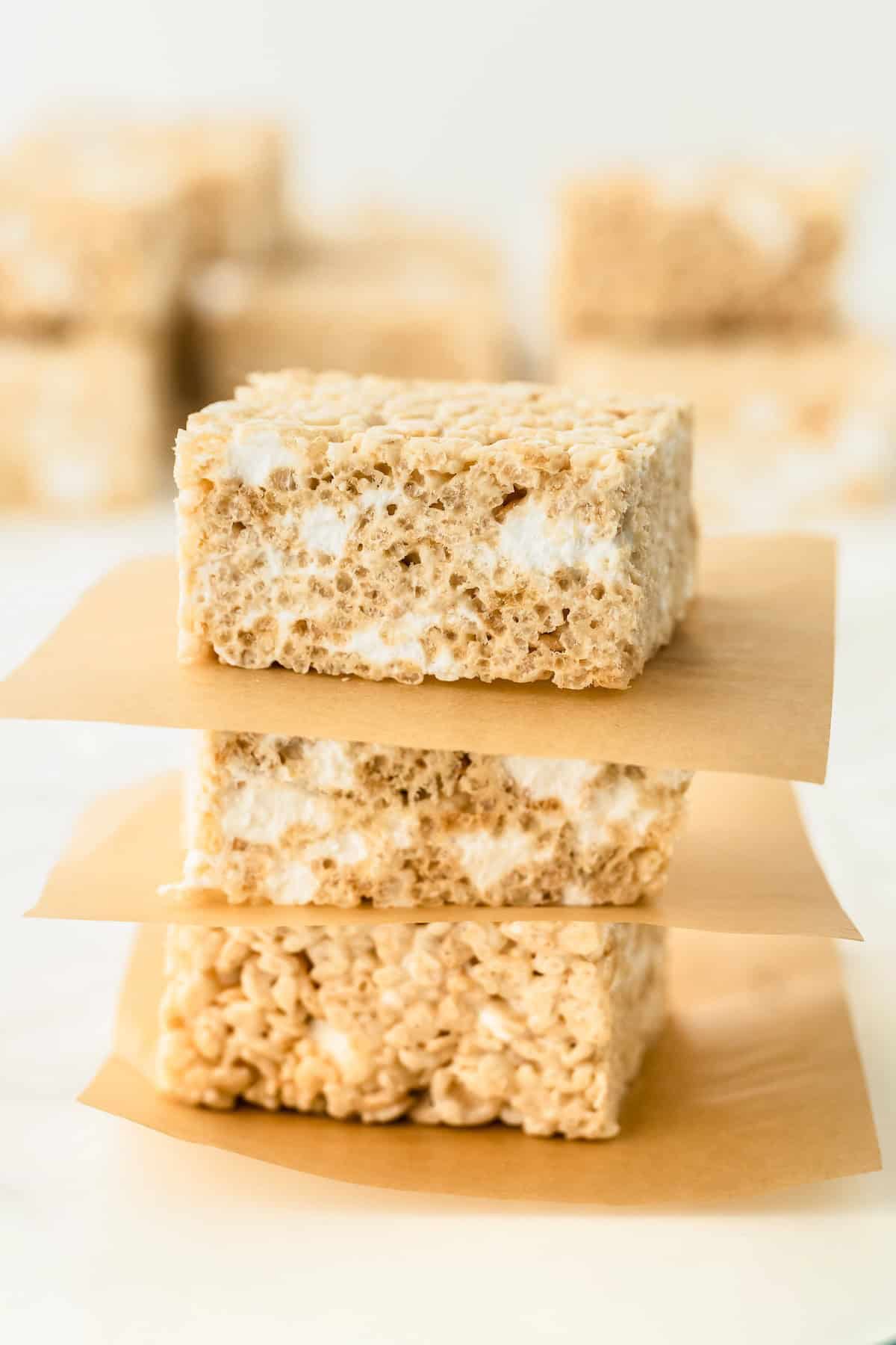 Three browned butter rice krispie treats with visible marshmallows are stacked on top of each other, each separated by a square piece of brown parchment paper. More treats are blurred in the background.