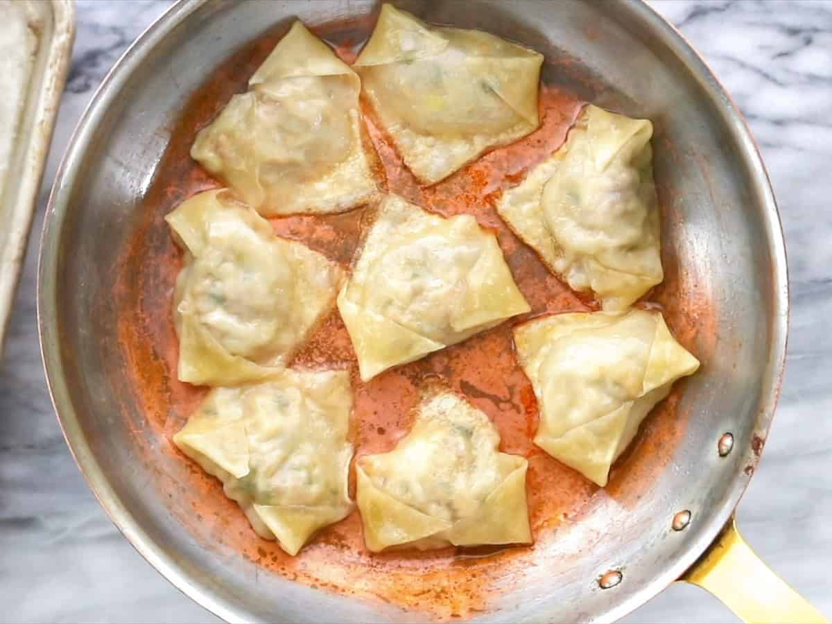 Potstickers are cooking in a stainless steel skillet with a red-tinted sauce. The dumplings are arranged in a circular pattern, and the skillet rests on a marble countertop.