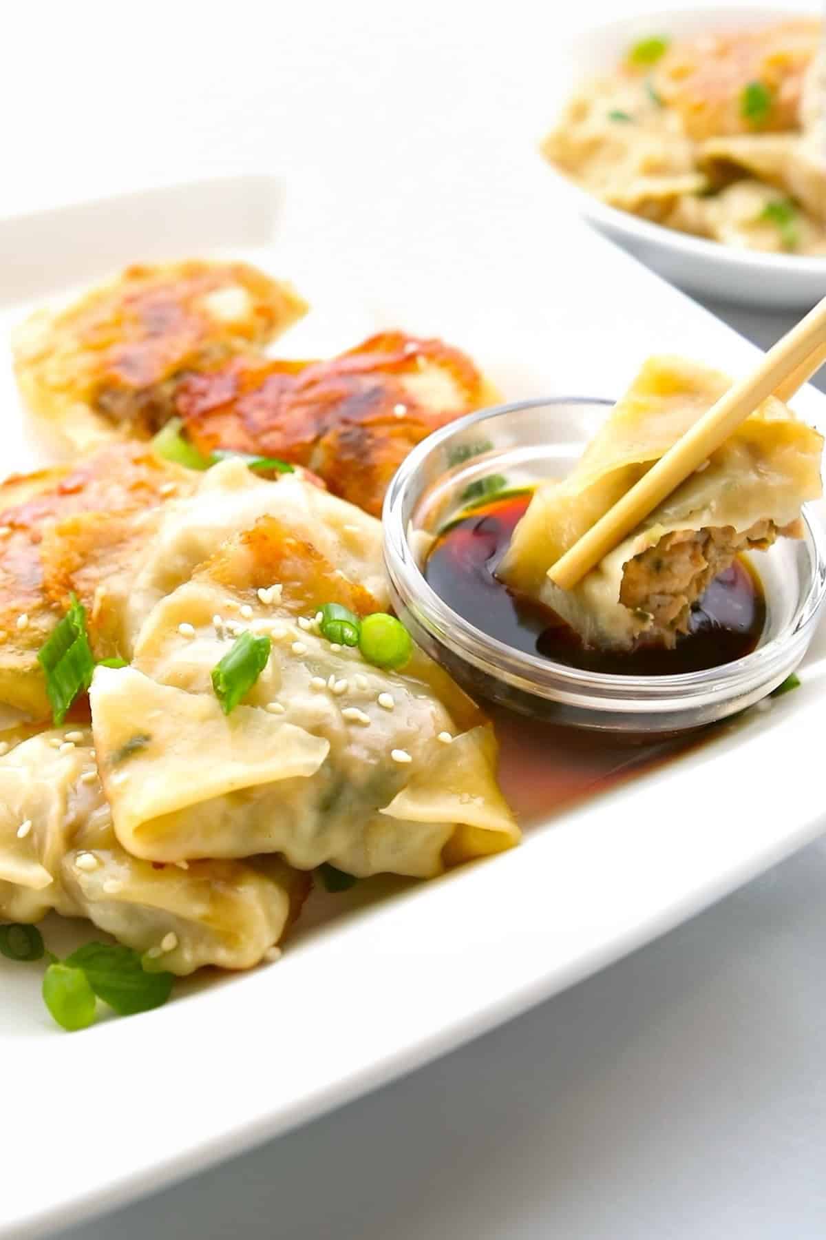A close-up of Chinese dumplings garnished with sesame seeds and green onions on a white plate. One potsticker is held by chopsticks and dipped into a small bowl of soy sauce.