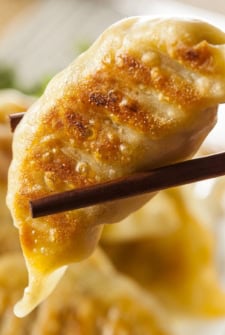 A close-up of chopsticks holding a golden-brown, pan-fried dumpling with crisp texture and visible grill marks. The background shows more dumplings out of focus.