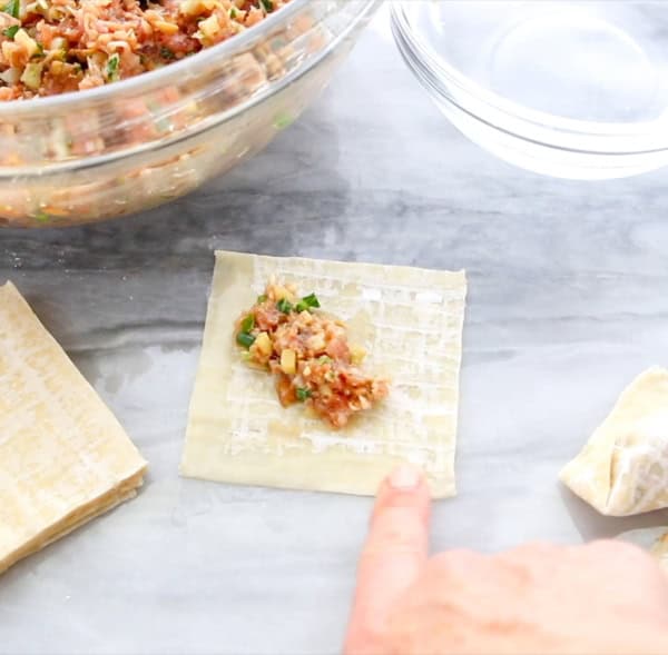 A hand points to a wonton wrapper with a spoonful of meat and vegetable filling on it, next to a stack of wrappers and a glass bowl of filling on a gray surface.