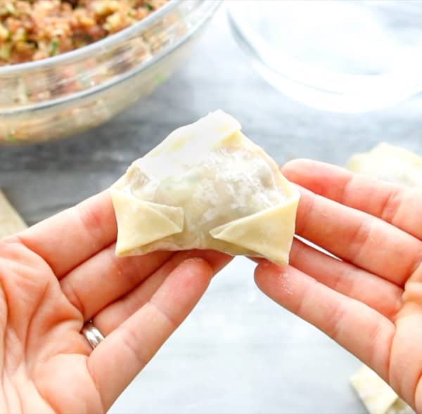 Two hands are holding a folded wonton wrapper stuffed with a meat filling. In the background, there is a glass bowl containing the remaining filling. The wonton wrapper is neatly folded and uncooked.